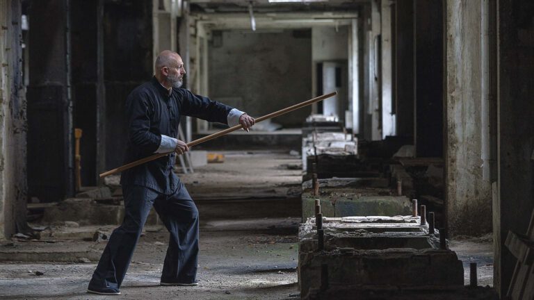 Ein Tai-Chi-Lehrer demonstriert eine Übung mit einem Langstock in einer ruhigen, ehemaligen Industriehalle mit natürlichem Lichteinfall.