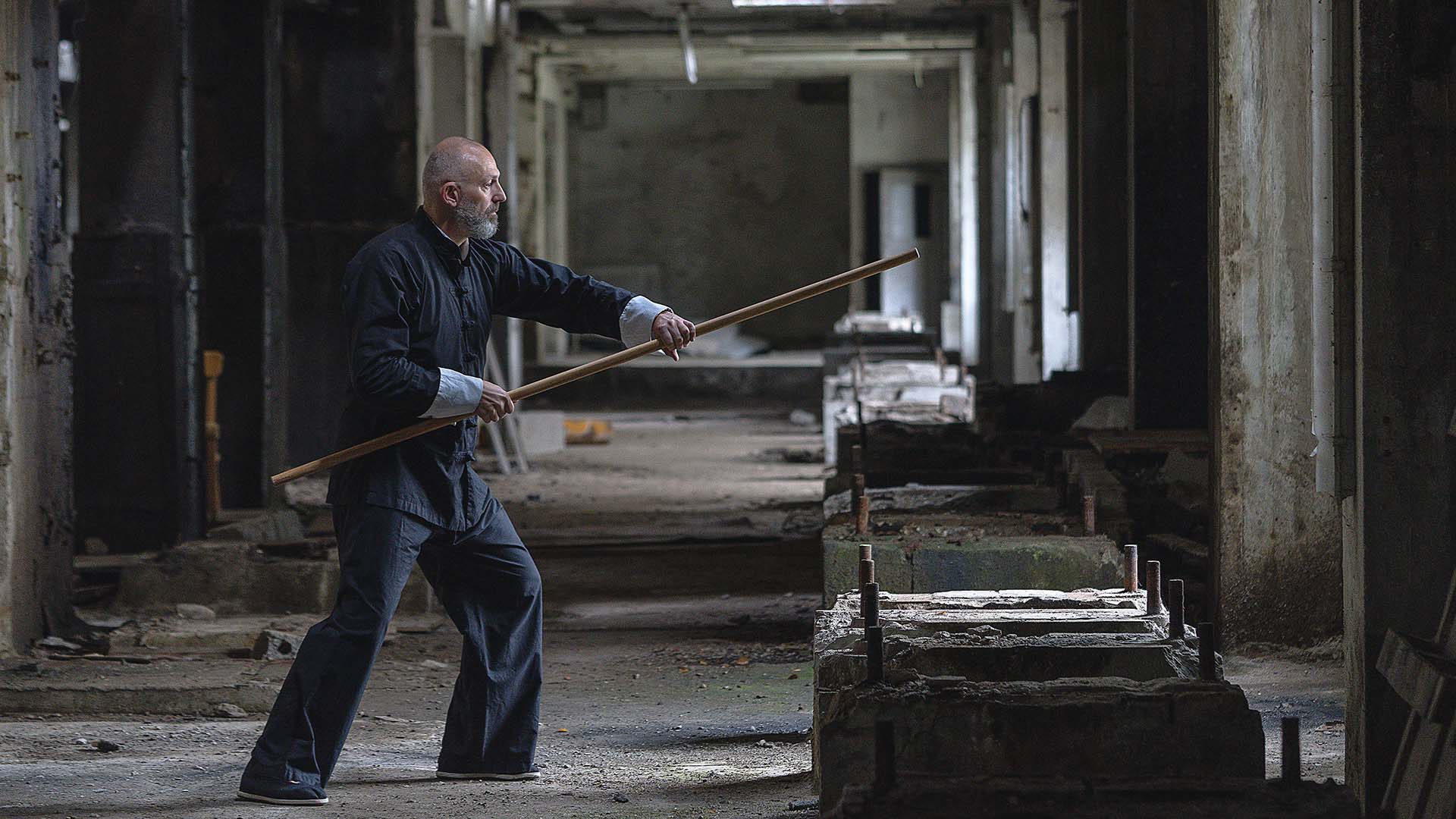 Ein Tai-Chi-Lehrer demonstriert eine Übung mit einem Langstock in einer ruhigen, ehemaligen Industriehalle mit natürlichem Lichteinfall.