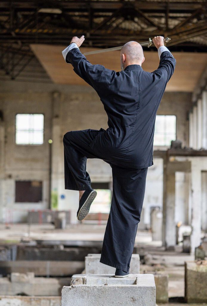 Tai-Chi-Schwertform auf einem Betonpodest in einer ehemaligen Industriehalle – Übung für Balance, Konzentration und innere Stabilität.