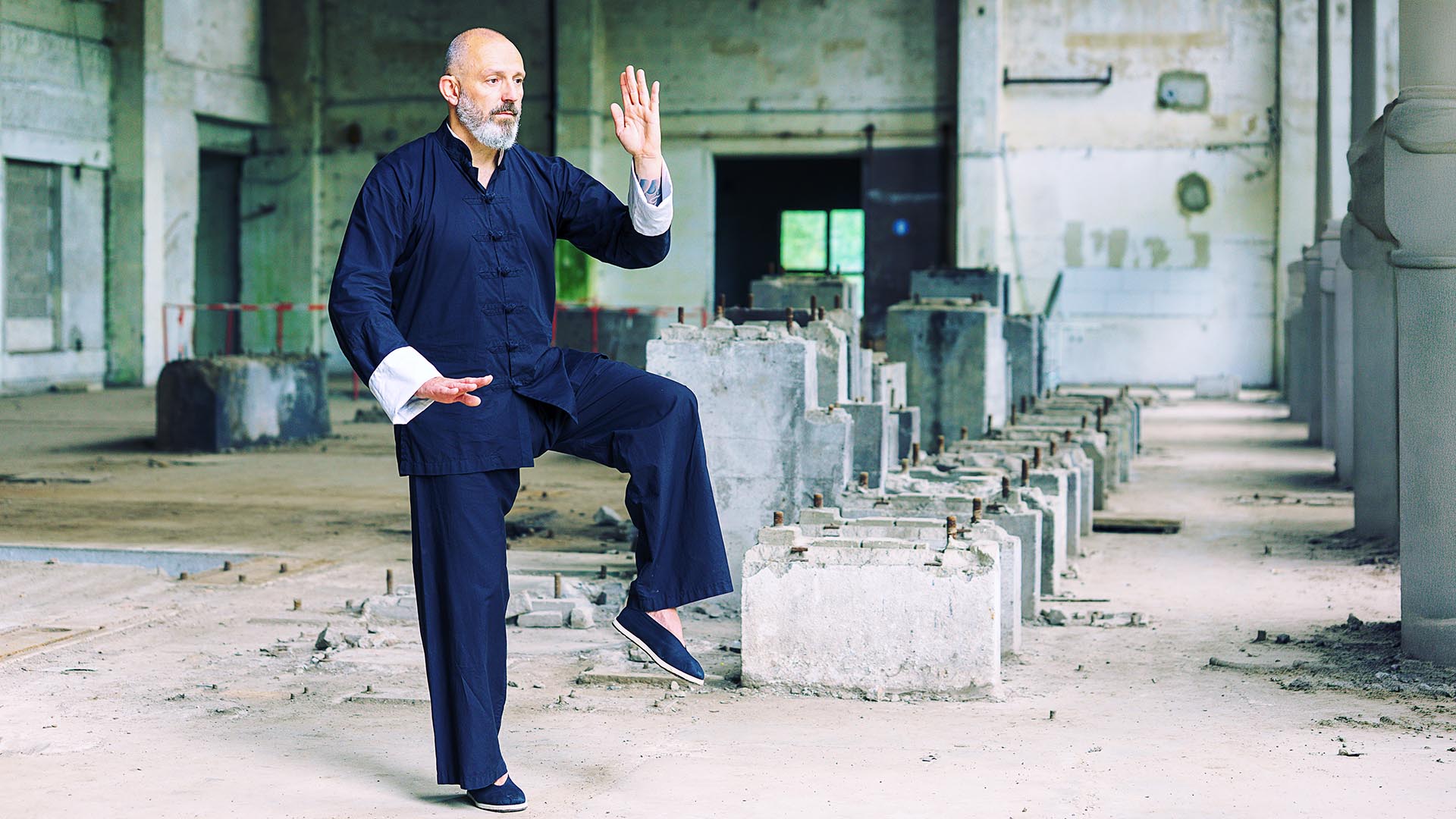 Ein Mann in traditioneller dunkler Kampfkunstkleidung steht in einer verlassenen Halle auf einem Bein und hebt eine Hand nach oben