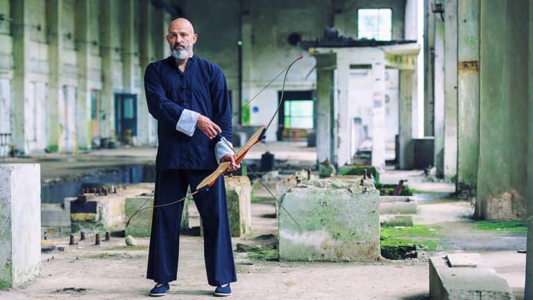 Georg Daun mit Bart in traditioneller Kleidung steht in einer verlassenen Industriehalle und hält einen Bogen in der Hand