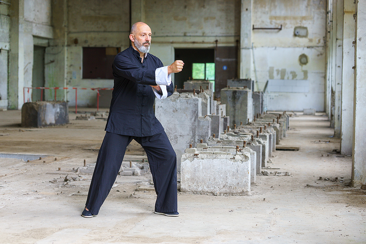 Georg Daun steht in traditioneller Tai chi chuan Kleidung in einer Pose in einer Halle eines Lost Places und trainiert.