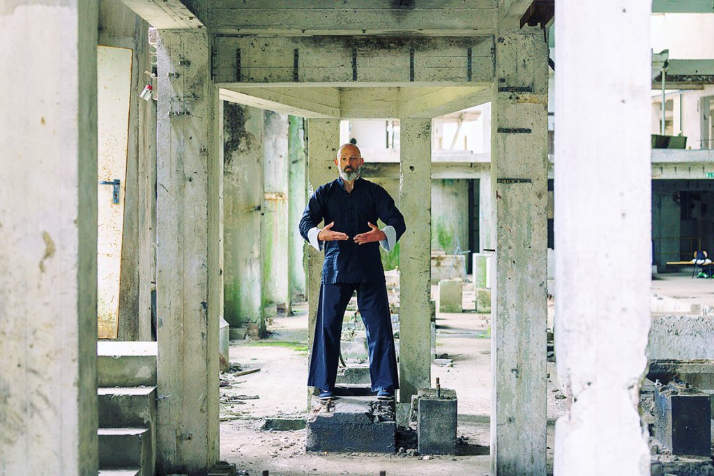Georg Daun, steht in traditioneller, dunkler Kampfkunstkleidung, in einer verlassenen Industrie Halle, einem Lost Place, aufrecht in einer Pose des Zhan Zhuang Qigong.