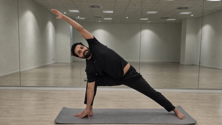 Yoga-Lehrer Neeraj in schwarzer Kleidung führt eine seitliche Dehnübung auf einer grauen Matte in einem hellen, spiegelnden Raum aus.